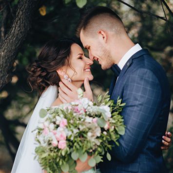 Bride and groom share an intimate moment beneath a tree at The Garden Venue wedding venue, with the groom gently holding the bride’s face and the bride smiling while holding a bouquet of pink and white flowers.