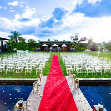 Outdoor ceremony setup at The Garden Venue wedding venue, featuring rows of white chairs arranged on a manicured lawn, a striking red carpet aisle over a reflective pond, and elegant floral arrangements under a vibrant blue sky.