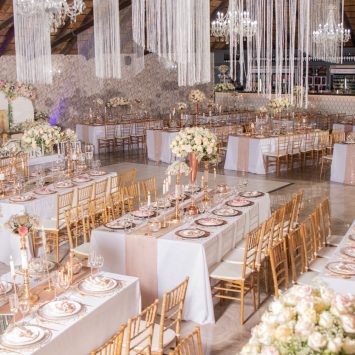 Luxurious indoor reception setup at The Garden Venue wedding venue, featuring long banquet tables with blush and gold accents, elegant floral centerpieces, gold chairs, and cascading ceiling drapes under a thatched roof and chandeliers.
