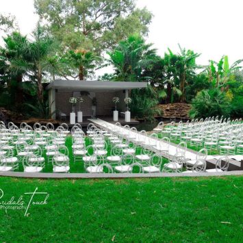 Outdoor ceremony setup at The Garden Venue wedding venue, showcasing a modern stage with white floral arrangements, surrounded by clear chairs arranged on lush green grass and a wooden walkway crossing over a tranquil water feature.
