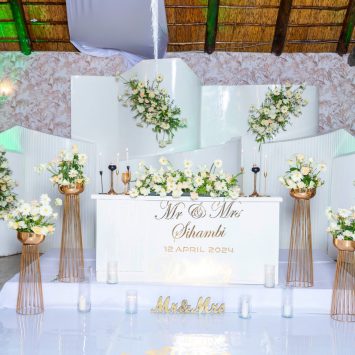 Elegant indoor wedding setup at The Garden Venue wedding venue, showcasing a modern white stage with gold accents, floral arrangements in white and yellow tones, candles, and a personalized sweetheart table for the bride and groom.