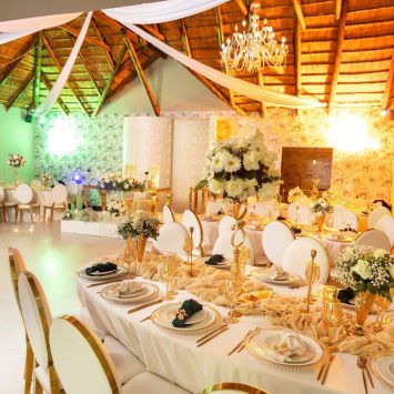 Indoor reception setup at The Garden Venue wedding venue, featuring elegant round and rectangular tables adorned with gold accents, white floral centerpieces, and chic white chairs under a thatched roof ceiling with chandeliers and draped fabric.